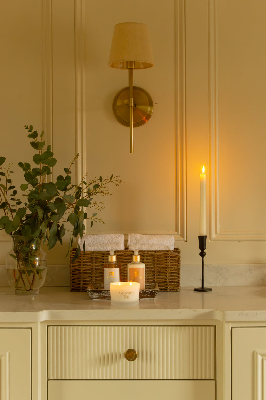 Decorative setting with candles, Bath and Body Wash & Lotion, a basket with towels, and a plant on a marble countertop with a wall-mounted lamp in the background.