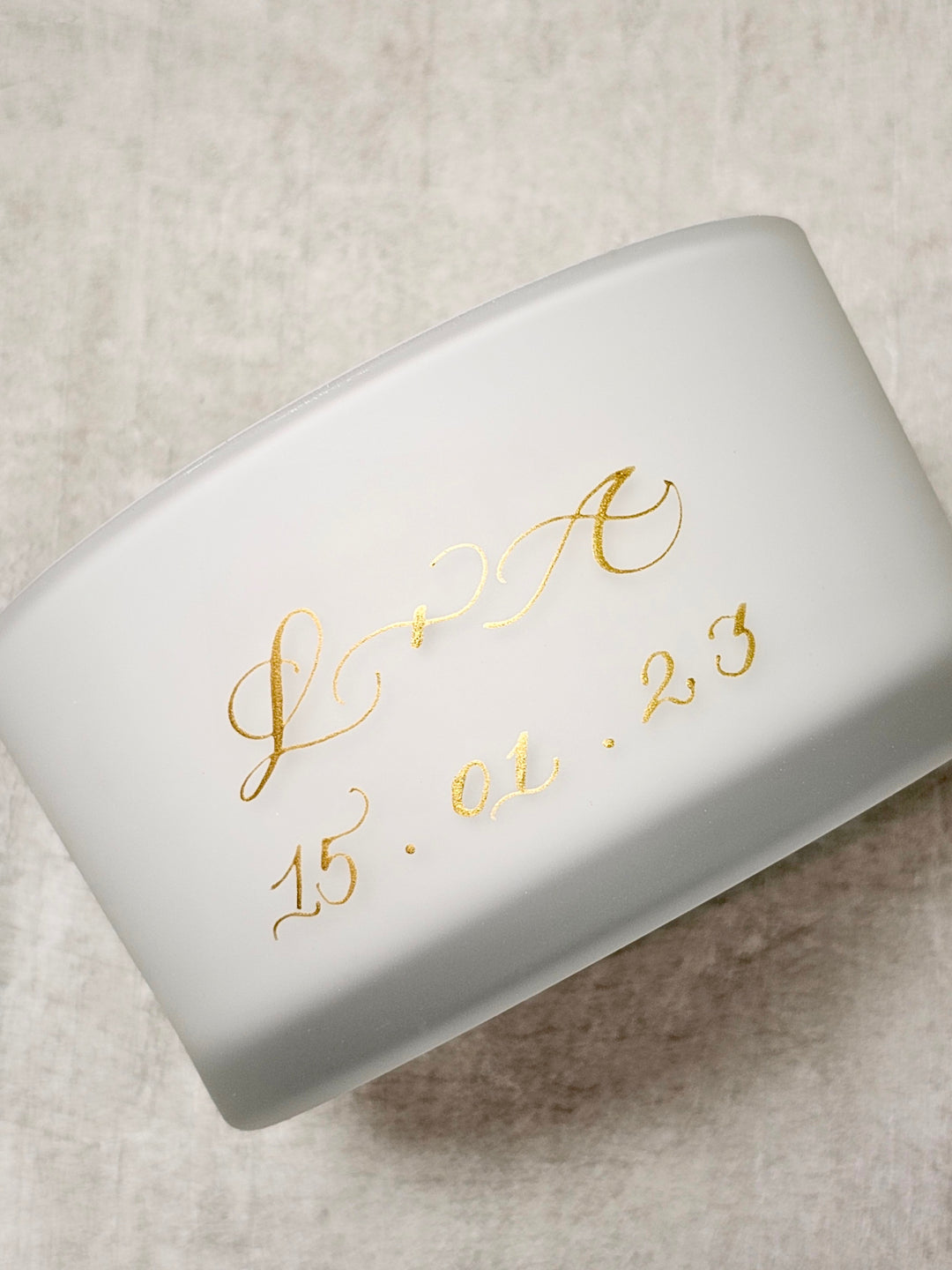 White candle with gold cursive text on a textured beige background.