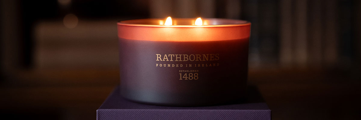 Rathbornes Seanchaí Dún classic 2 wick candle lit in plum glass, standing on its aubergine purple gift box in a library