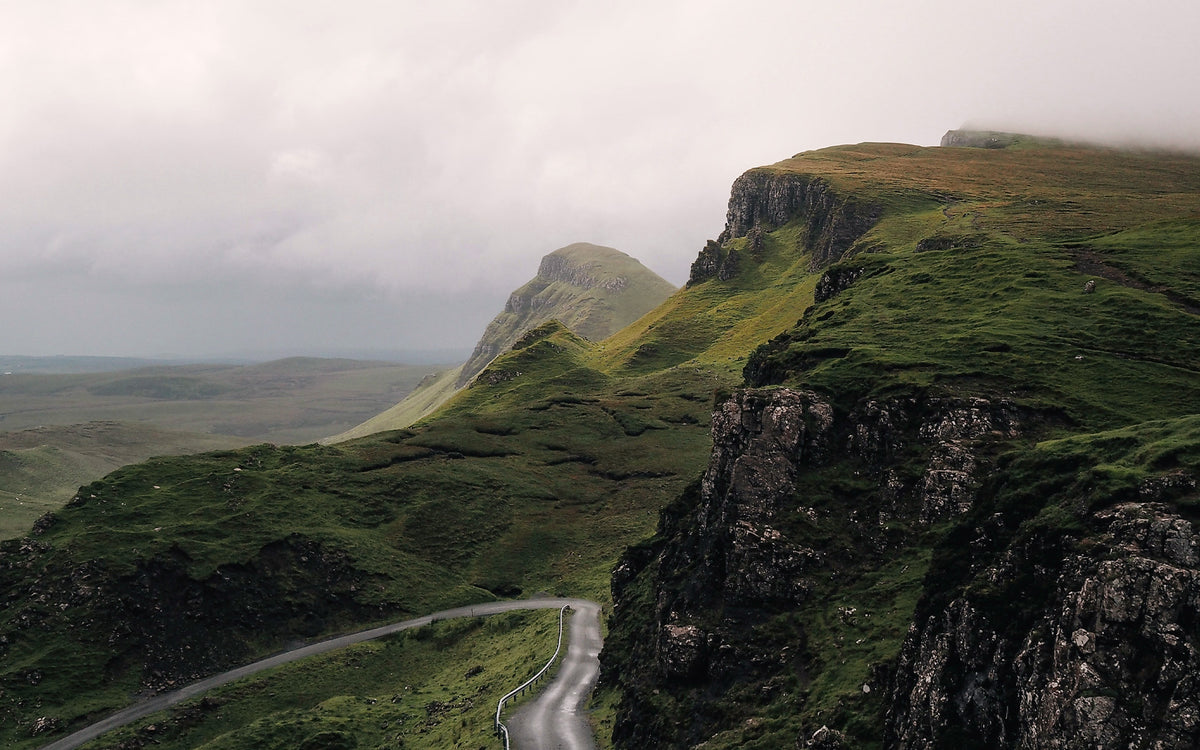 Misty green hills and rocky cliffs above a winding single-track road under a low grey autumn sky