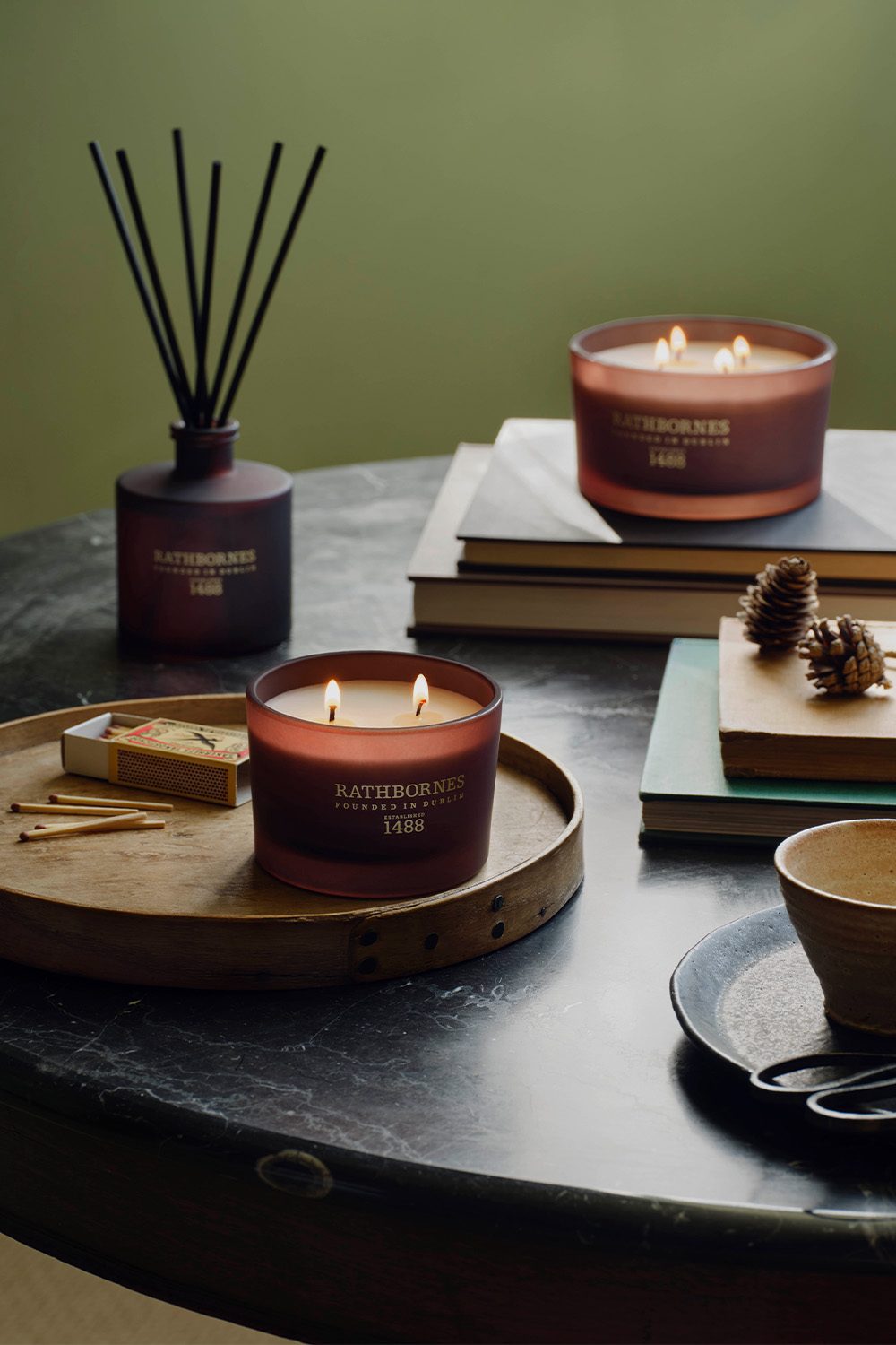Rathbornes scented candles and reed diffuser on a marble table with matches and pine cones
