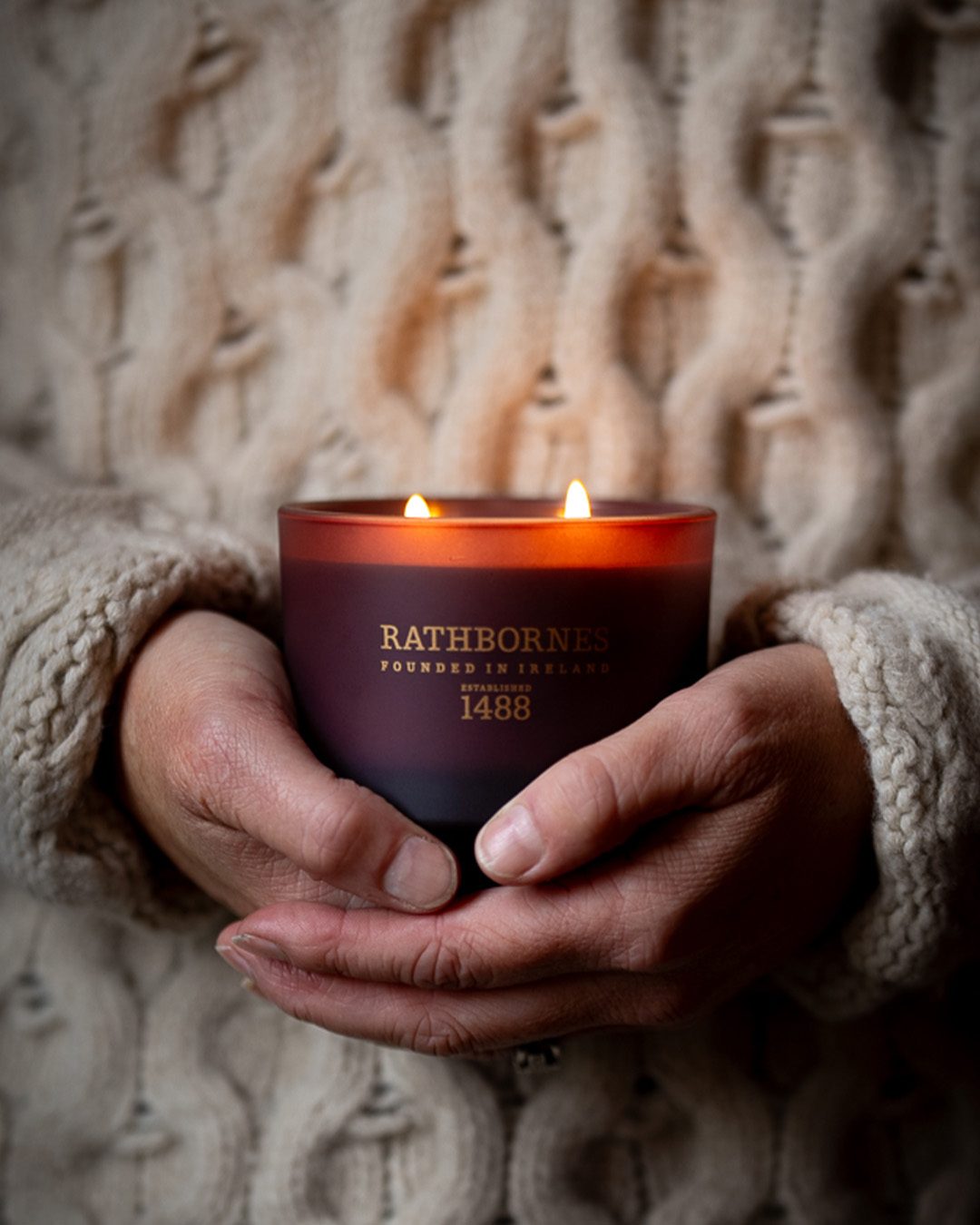 Hands in a cream cable-knit jumper holding a lit Rathbornes Seanchaí Dún scented candle in plum glass